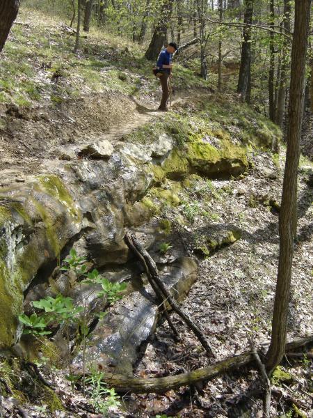 A hiker on a trail in a wooded area, positioned near a rocky outcrop with moss, while a small stream flows alongside. The scene captures greenery and trees in the background, showcasing a serene outdoor environment. Brown County Park mountain bike trail.