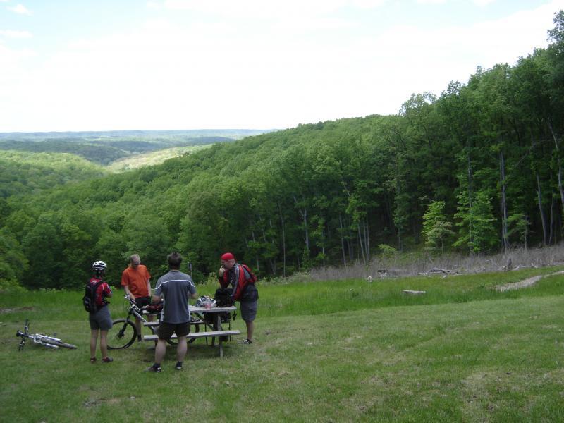 Four individuals gather around a picnic table in a lush, green outdoor setting, with a scenic view of rolling hills in the background. Two people are wearing helmets, indicating they have been biking, while others are engaged in conversation and preparing food on the table. The scene is bright and sunny, showcasing a vibrant natural landscape. Brown County Park mountain bike trail.