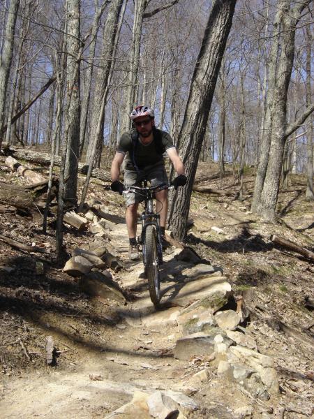 A mountain biker navigating a rocky trail through a forested area, with trees in the background and a clear blue sky overhead. The cyclist is wearing a helmet and gloves, riding a mountain bike along a winding path made of stones. Brown County Park mountain bike trail.