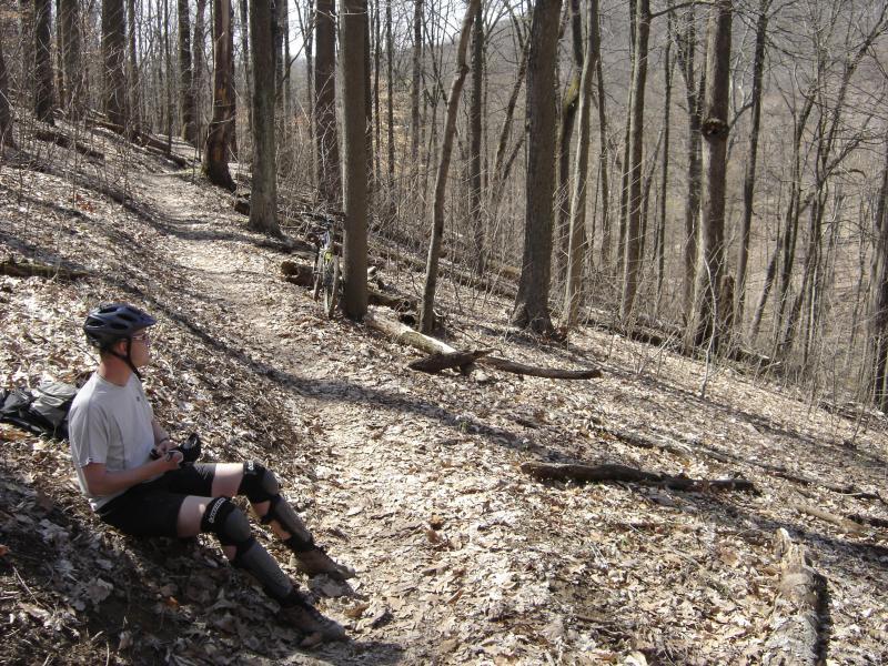 A person sitting on the ground along a forest trail, wearing a helmet and kneepads, with a bicycle nearby. The scene features bare trees and a leaf-covered path, indicating early spring or late winter conditions. Brown County Park mountain bike trail.