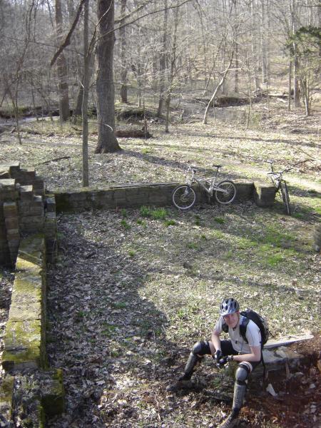 A cyclist resting on a stone foundation in a wooded area. Two mountain bikes are parked nearby on the forest floor, surrounded by fallen leaves and trees. The scene captures the tranquility of nature with sunlight filtering through the branches. Brown County Park mountain bike trail.