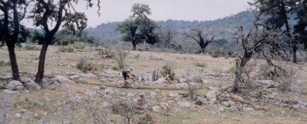 A cyclist riding on a narrow path through a rocky, sparsely vegetated landscape, surrounded by trees and hills in the background. The scene is set in a remote, natural area with an overcast sky. Flat Rock Ranch mountain bike trail.