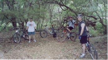 Three mountain bikers stand in a wooded area surrounded by trees, with their bicycles parked nearby. The ground is covered in leaves, and the lighting suggests a sunny day. The cyclists are wearing casual athletic clothing and helmets, and they appear to be taking a break or posing for the photo. McAllister Park mountain bike trail.