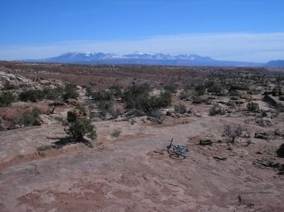 A wide, panoramic view of a rugged, arid landscape featuring rocky terrain and sparse vegetation, with distant snow-capped mountains under a clear blue sky. Sovereign Single Track mountain bike trail.