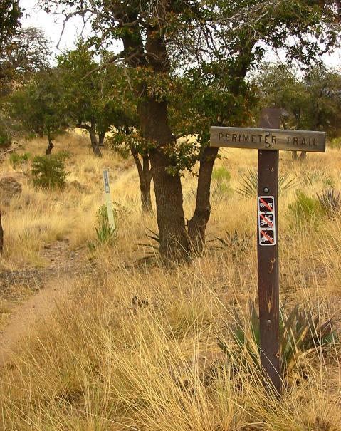 A wooden trail sign labeled "Perimeter Trail" stands in a grassy area with sparse trees. In the foreground, a narrow dirt path leads away from the sign. Another sign, partially visible, is located further down the trail, and there are several warning symbols displayed on the main sign. The landscape features golden grasses and scattered vegetation. Perimeter Trail mountain bike trail.