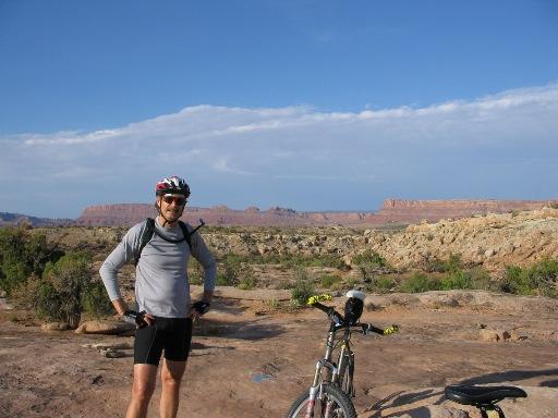 A cyclist wearing a helmet and athletic clothing stands confidently beside a mountain bike on a rocky terrain, with desert landscapes and blue skies in the background. Sovereign Single Track mountain bike trail.