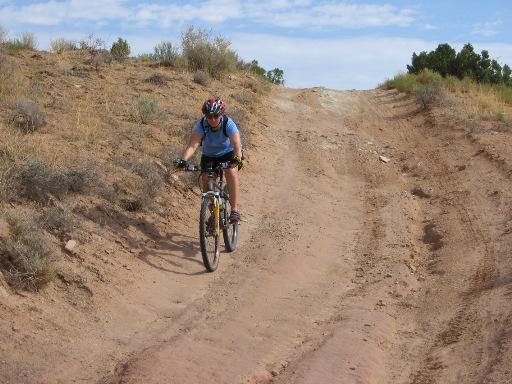 A mountain biker navigating a dirt trail with rocky terrain, surrounded by sparse vegetation and hills under a partly cloudy sky. Sovereign Single Track mountain bike trail.