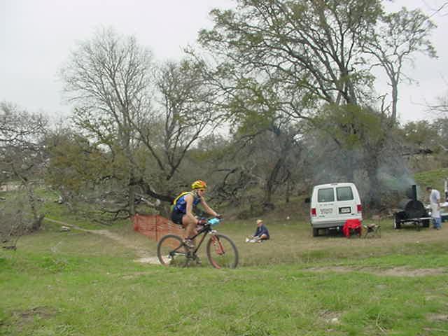A cyclist wearing a helmet and sports gear rides a mountain bike along a dirt path in a grassy area. In the background, a white van is parked near some trees, and a small group of people is gathered nearby, one of whom is seated. Smoke rises from a grilling area, indicating a casual outdoor gathering. The scene is set on a cloudy day, highlighting a blend of nature and recreational activity. Flat Rock Ranch mountain bike trail.
