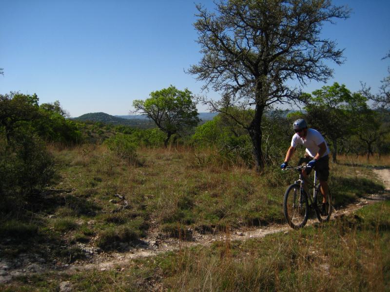 A person riding a mountain bike on a dirt trail surrounded by green trees and grassy terrain under a clear blue sky. Flat Rock Ranch mountain bike trail.