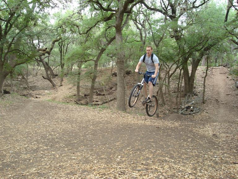 A person riding a mountain bike is performing a jump on a dirt trail surrounded by trees. The ground is covered in fallen leaves, and the scene captures a moment of action in a natural outdoor setting. McAllister Park mountain bike trail.