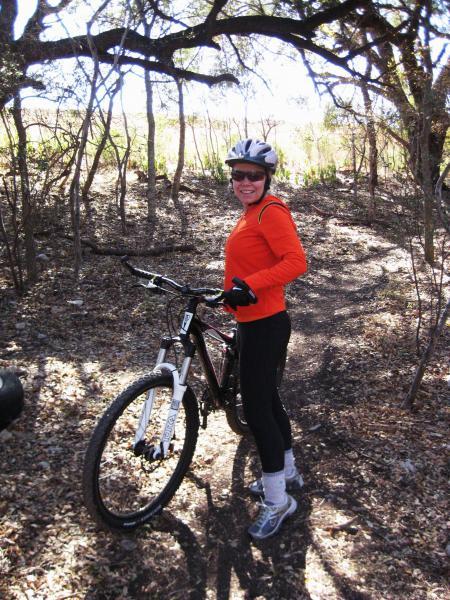A smiling person wearing a bright orange long-sleeve shirt, gloves, and a helmet stands next to a mountain bike on a dirt trail surrounded by trees and dry leaves. The background features a sunny, natural setting. McAllister Park mountain bike trail.