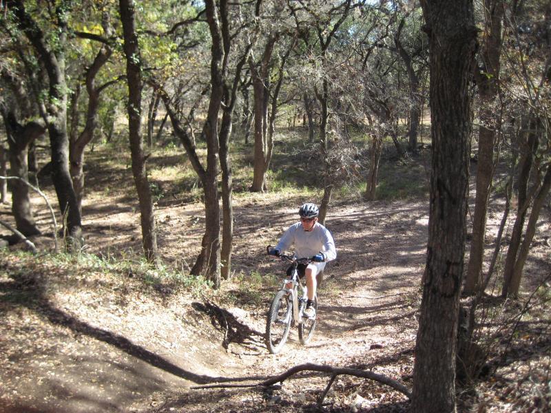 A person riding a mountain bike on a dirt trail through a wooded area, surrounded by trees and natural foliage. McAllister Park mountain bike trail.