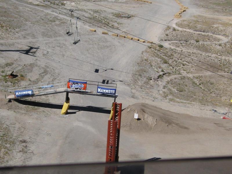 A scenic view of a ski resort area featuring a ski lift in the background, with a wooden platform marked with signs at the forefront. The ground is primarily dirt and gravel, indicating off-season or summer conditions, and there's a small jump or mound visible. The environment appears rugged and mountainous. Mammoth Mountain Resort mountain bike trail.