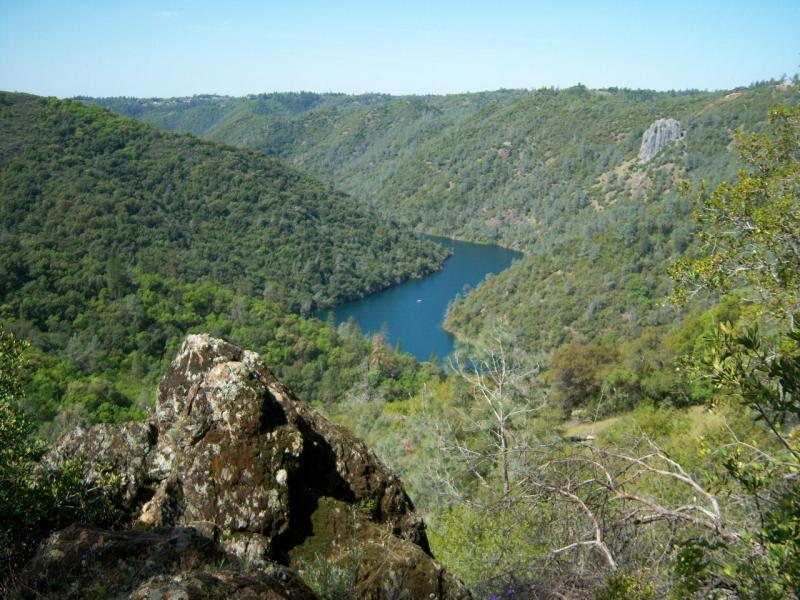 A scenic view of a lush green valley with a winding blue lake surrounded by hills. Large, rocky foreground in the lower left corner adds depth to the landscape, while vibrant foliage covers the hillsides, showcasing a bright blue sky above. Clementine / Forresthill Connector Trail mountain bike trail.