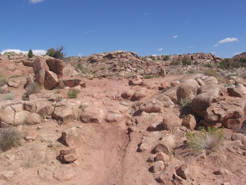 A rocky dirt path winding through a dry landscape with scattered boulders and sparse vegetation under a blue sky with a few clouds. Sovereign Single Track mountain bike trail.
