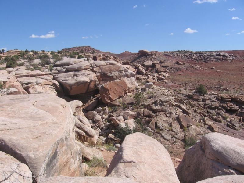 Rocky landscape with large boulders and scattered shrubs under a bright blue sky with a few clouds. The terrain features a variety of rock formations and layers of soil, showcasing the natural beauty of a desert environment. Sovereign Single Track mountain bike trail.
