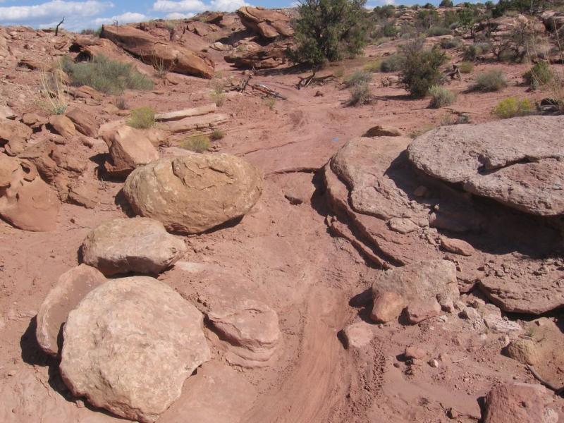 A rocky landscape featuring various sizes of boulders on sandy terrain, with sparse vegetation visible. The scene includes a winding dirt path leading through the rocks under a blue sky with scattered clouds. Sovereign Single Track mountain bike trail.