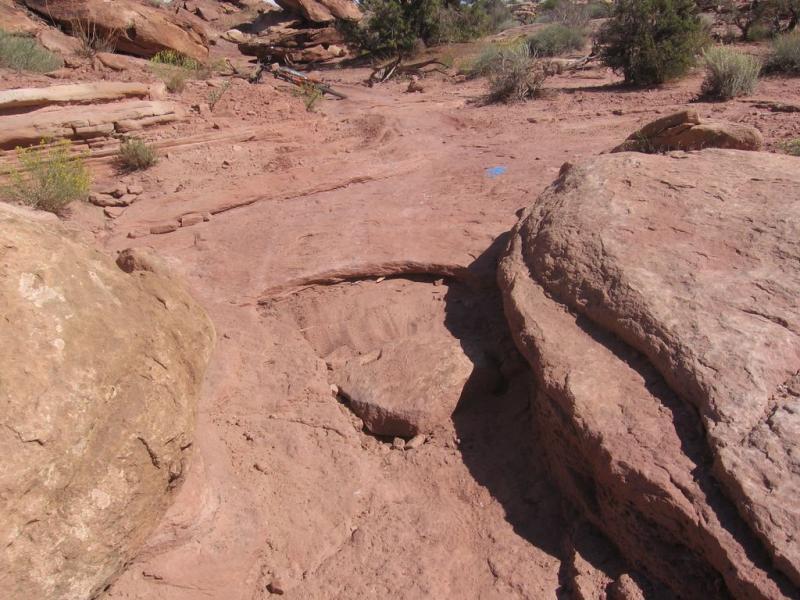 A rocky, sandy terrain featuring large boulders and sparse vegetation. The ground is uneven with visible paths and signs of erosion, set in a natural outdoor environment. Sovereign Single Track mountain bike trail.