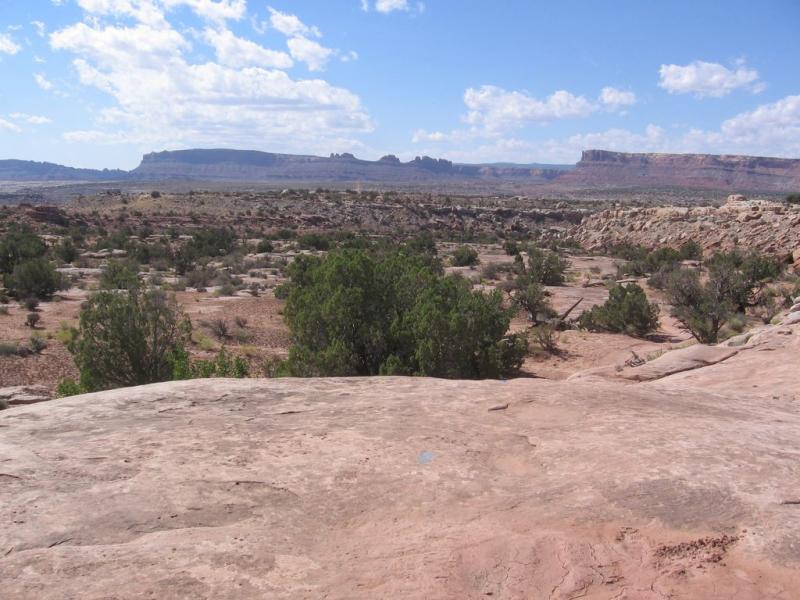 A panoramic view of a rugged landscape featuring rocky formations, sparse vegetation, and a broad sky with scattered clouds. The foreground includes a flat rocky surface, while the background showcases distant mesas and cliffs under bright daylight. Sovereign Single Track mountain bike trail.