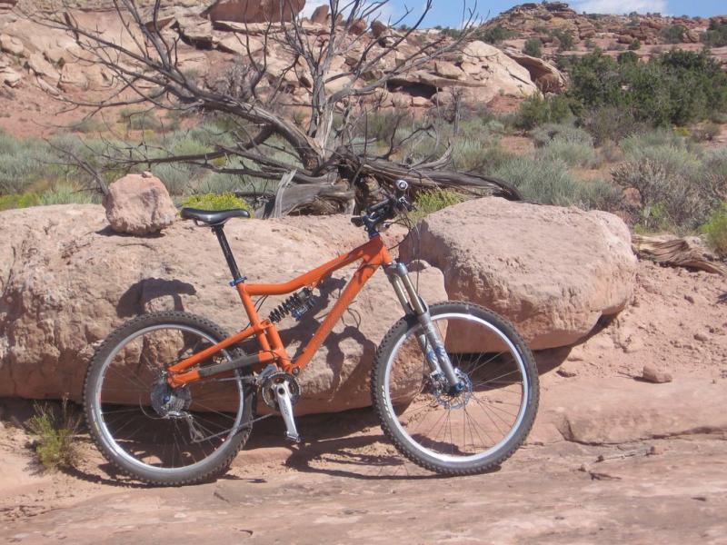A bright orange mountain bike is parked next to a large rock in a natural setting, with sparse vegetation and rocky terrain in the background. The scene features arid landscape elements, including a distant rocky outcrop and a bare tree in the background under a clear blue sky. Sovereign Single Track mountain bike trail.