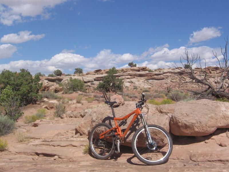 A mountain bike with an orange frame parked on rocky terrain, surrounded by sparse vegetation and a blue sky dotted with clouds. Sovereign Single Track mountain bike trail.