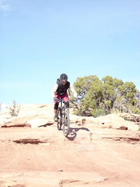 A mountain biker navigating a rocky trail under a clear blue sky, with rugged terrain and sparse vegetation visible in the background. The cyclist is wearing a helmet and riding gear, focused on balancing the bike as they descend the rocky surface. Sovereign Single Track mountain bike trail.