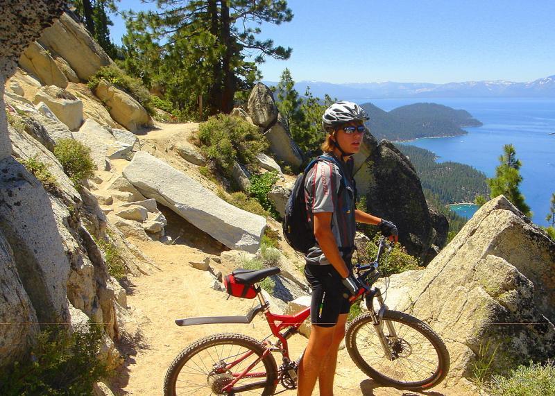 A mountain biker stands next to a red mountain bike on a rocky trail surrounded by trees, overlooking a scenic view of a lake and mountains in the distance. The sky is clear and blue, indicating a sunny day. Tahoe Rim Trail: Tahoe Meadows to Tunnel Creek Road / Flume Trail mountain bike trail.