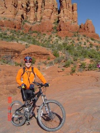 A person in an orange cycling jacket and helmet is standing next to a mountain bike on rocky terrain. Behind them, red rock formations and greenery create a natural landscape under a clear blue sky. Broken Arrow Trail / Chicken Point mountain bike trail.
