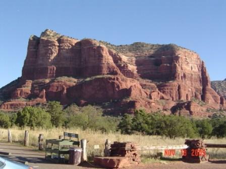 A large red rock formation rises prominently against a clear blue sky, surrounded by green vegetation and a grassy field. In the foreground, there's a bench and some stacked stones, indicating a visitor area. Broken Arrow Trail / Chicken Point mountain bike trail.
