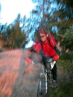 A mountain biker in a red shirt and black shorts navigating over rocky terrain, with a blurred effect suggesting motion. The background features trees and a dusky sky, adding to the sense of outdoor adventure. Horsetooth Mountain Park mountain bike trail.