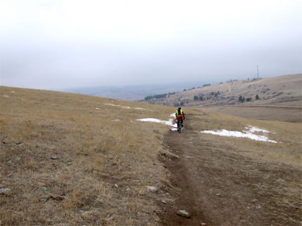 A cyclist riding on a dirt path through a grassy, hilly landscape under an overcast sky. The terrain appears to be slightly snowy in patches, and the cyclist is wearing a reflective vest and carrying a backpack. Marshall Mesa Area mountain bike trail.