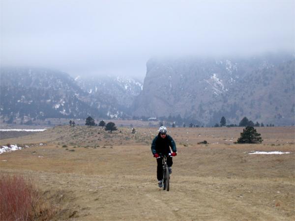 A cyclist riding on a dirt path through a rural landscape, with mountains shrouded in clouds in the background. The scene features a mix of open terrain and sparse vegetation, suggesting a cool, misty day. Marshall Mesa Area mountain bike trail.