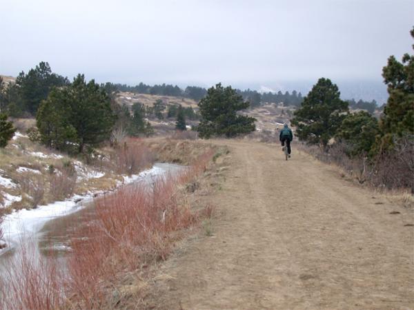 A solitary person walking along a dirt path beside a stream, surrounded by sparse vegetation and pine trees. The landscape features rolling hills in the background under a cloudy sky. Some areas of snow are visible on the ground, indicating a chilly atmosphere. Marshall Mesa Area mountain bike trail.