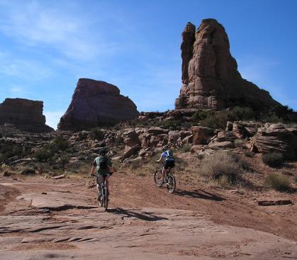 Two mountain bikers riding along a rocky trail in a desert landscape, with towering rock formations and a clear blue sky in the background. Amasa Back Trail mountain bike trail.