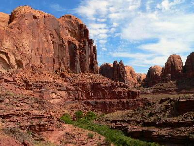 A scenic view of rugged red rock formations under a clear blue sky, showcasing a dramatic landscape with cliffs and canyons. Lush green vegetation is visible in the foreground, contrasting with the warm tones of the rocky terrain. Amasa Back Trail mountain bike trail.