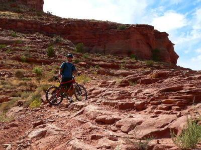 A person standing beside a mountain bike on a rocky, red dirt trail, surrounded by arid landscape and cliffs, under a blue sky with scattered clouds. Amasa Back Trail mountain bike trail.