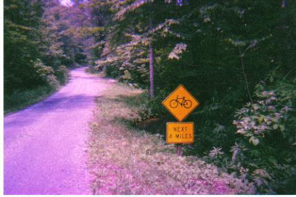 A gravel road winding through a wooded area with a yellow bicycle warning sign indicating that bicycle routes continue for the next four miles. Bushwhacker Trail mountain bike trail.
