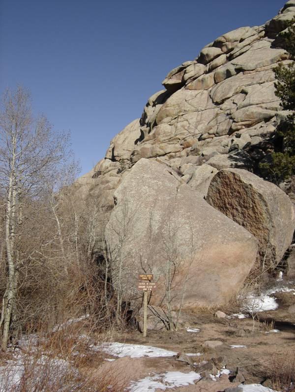 A large boulder stands prominently at the base of a rocky cliff, surrounded by sparse vegetation and patches of snow. A wooden sign next to the boulder is partially visible. The sky is clear and blue, indicating a bright sunny day. Vedauwoo mountain bike trail.