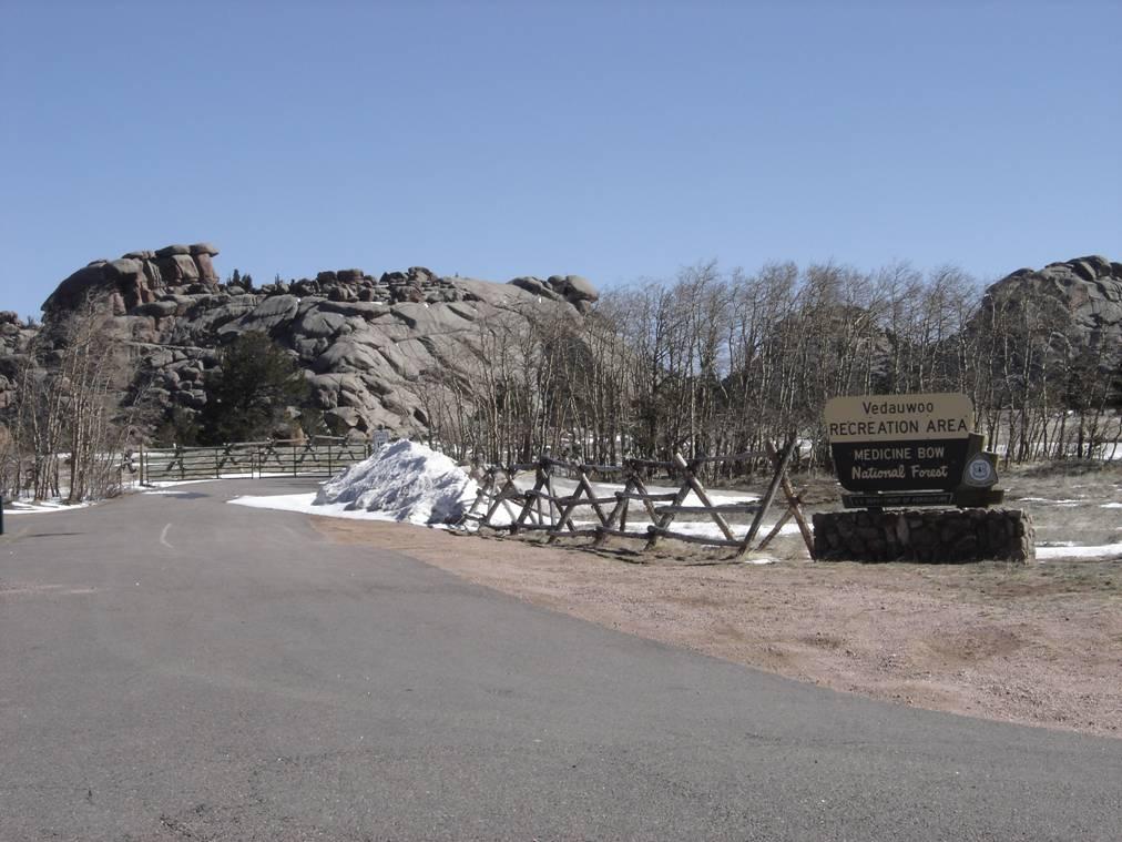 A scenic view of the Vedauwoo Recreation Area sign, surrounded by rocky formations and sparse trees, with a clear blue sky above. There is a snow pile on the side of the road, indicating early spring conditions. A wooden fence lines the area, providing a rustic touch to the natural landscape. Vedauwoo mountain bike trail.
