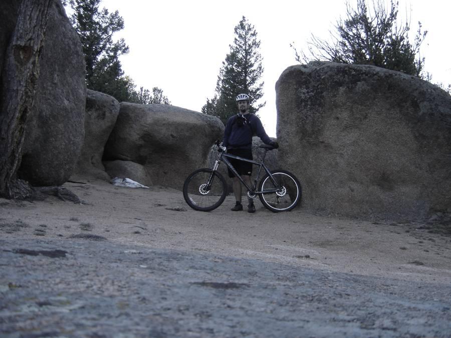 A person in a cycling helmet stands beside a mountain bike on a dirt path surrounded by large boulders and pine trees. The scene is set in a natural outdoor environment, with an overcast sky. Vedauwoo mountain bike trail.