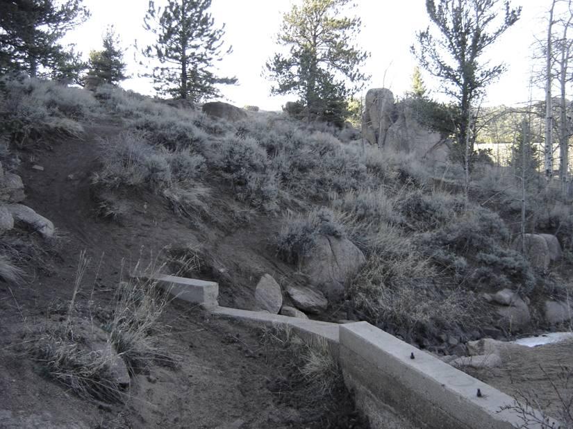 A rocky hillside with sparse vegetation, including tall grasses and shrubs. In the foreground, there is a concrete structure partially protruding from the ground, leading up to a steep path that ascends the hill. Surrounding trees are visible in the background, indicating a natural, outdoor setting. Vedauwoo mountain bike trail.