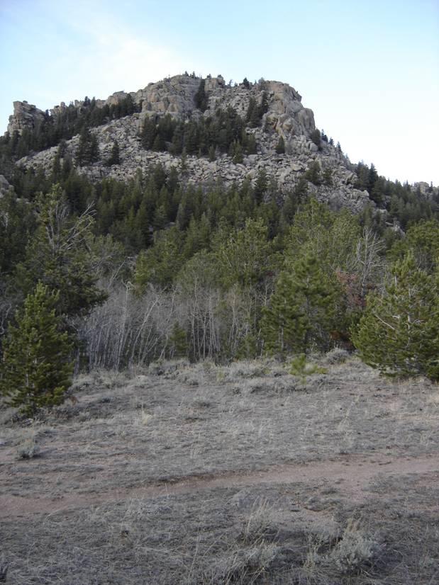 A rocky mountain peak rises above a forest of evergreen trees, with a trail meandering through the foreground. The landscape is earthy and rugged, showcasing a mix of rocky outcrops and patches of grass. The sky above is clear with hints of blue. Vedauwoo mountain bike trail.