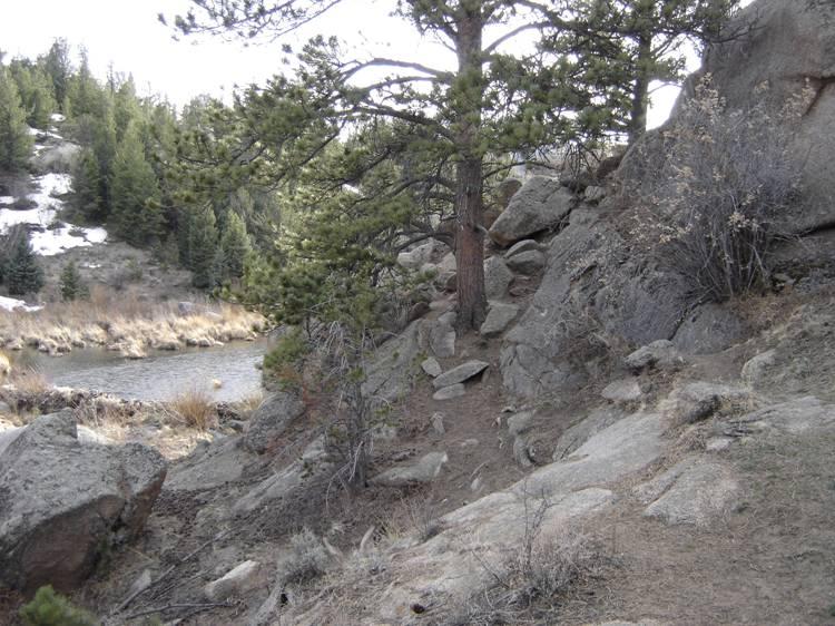 A rocky hillside with scattered boulders and sparse vegetation, including pine trees. In the background, a small river winds through a grassy area, with patches of snow visible on higher ground. The scene depicts a natural landscape in an outdoor setting. Vedauwoo mountain bike trail.