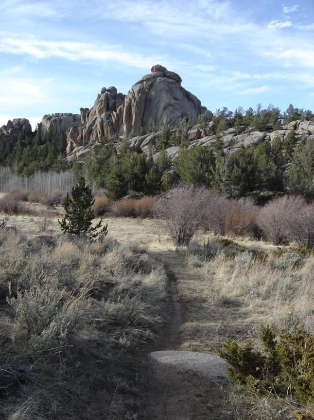 A scenic landscape featuring a winding dirt path leading through grassy terrain, surrounded by shrubs and sparse trees. In the background, large rock formations rise against a blue sky with scattered clouds, showcasing a natural, rugged environment. Vedauwoo mountain bike trail.