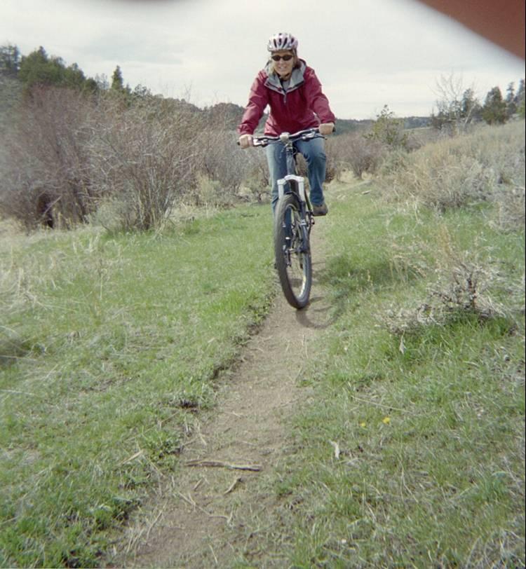 A person riding a mountain bike on a narrow dirt path, surrounded by green grass and shrubs, under a cloudy sky. The rider is wearing a red jacket and a helmet, enjoying the outdoor activity. Vedauwoo mountain bike trail.