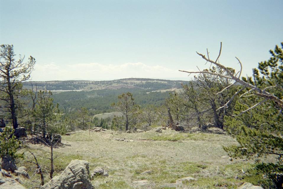 A panoramic view of a mountainous landscape with green trees and rocky outcrops. The foreground features a mix of grassy areas and scattered rocks, while the background showcases rolling hills and a clear blue sky with a few clouds. Sunlight illuminates the scene, highlighting the natural beauty of the area. Happy Jack Recreation Area mountain bike trail.