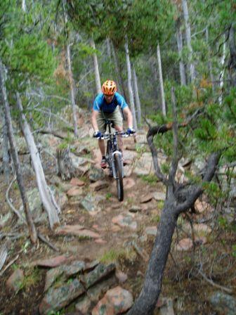 A mountain biker navigating a rocky trail in a forested area, wearing a helmet and riding downhill among trees and boulders. Happy Jack Recreation Area mountain bike trail.