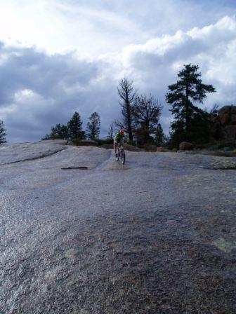 A mountain biker riding on a smooth, rocky surface under a cloudy sky, surrounded by tall trees and rocky outcrops. Vedauwoo mountain bike trail.