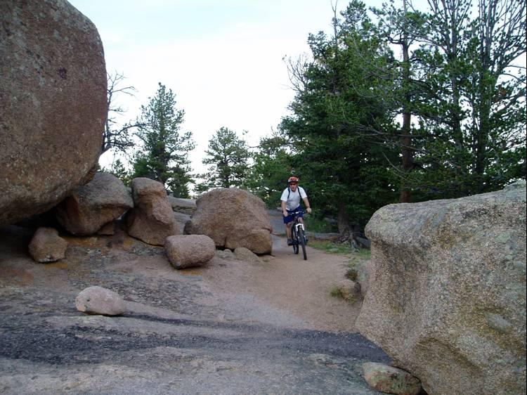 A person riding a mountain bike on a dirt trail surrounded by large rocks and trees in a natural outdoor setting. The cyclist is wearing a helmet and appears to be enjoying their ride through a rugged terrain. Vedauwoo mountain bike trail.