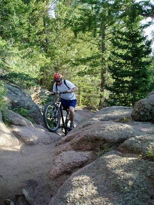 A mountain biker navigating a rocky trail in a forested area, carrying their bike over uneven terrain. Dense trees and greenery surround the path, showcasing a natural outdoor environment. The biker is wearing a helmet and casual sportswear. Vedauwoo mountain bike trail.